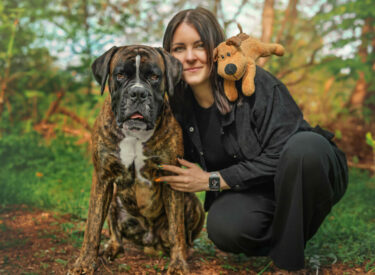 gemeinsames Fotoshooting mit Hund Boxer Baxter mit seiner Besitzerin Kopf an Kopf in der Hocke im Wald mit seinem Lieblingskuscheltier auf der Schulter